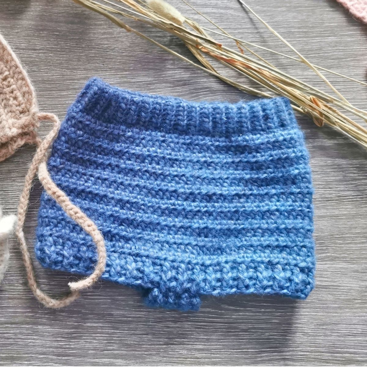 Blue knitted baby bloomers on a wooden surface with dried plants.